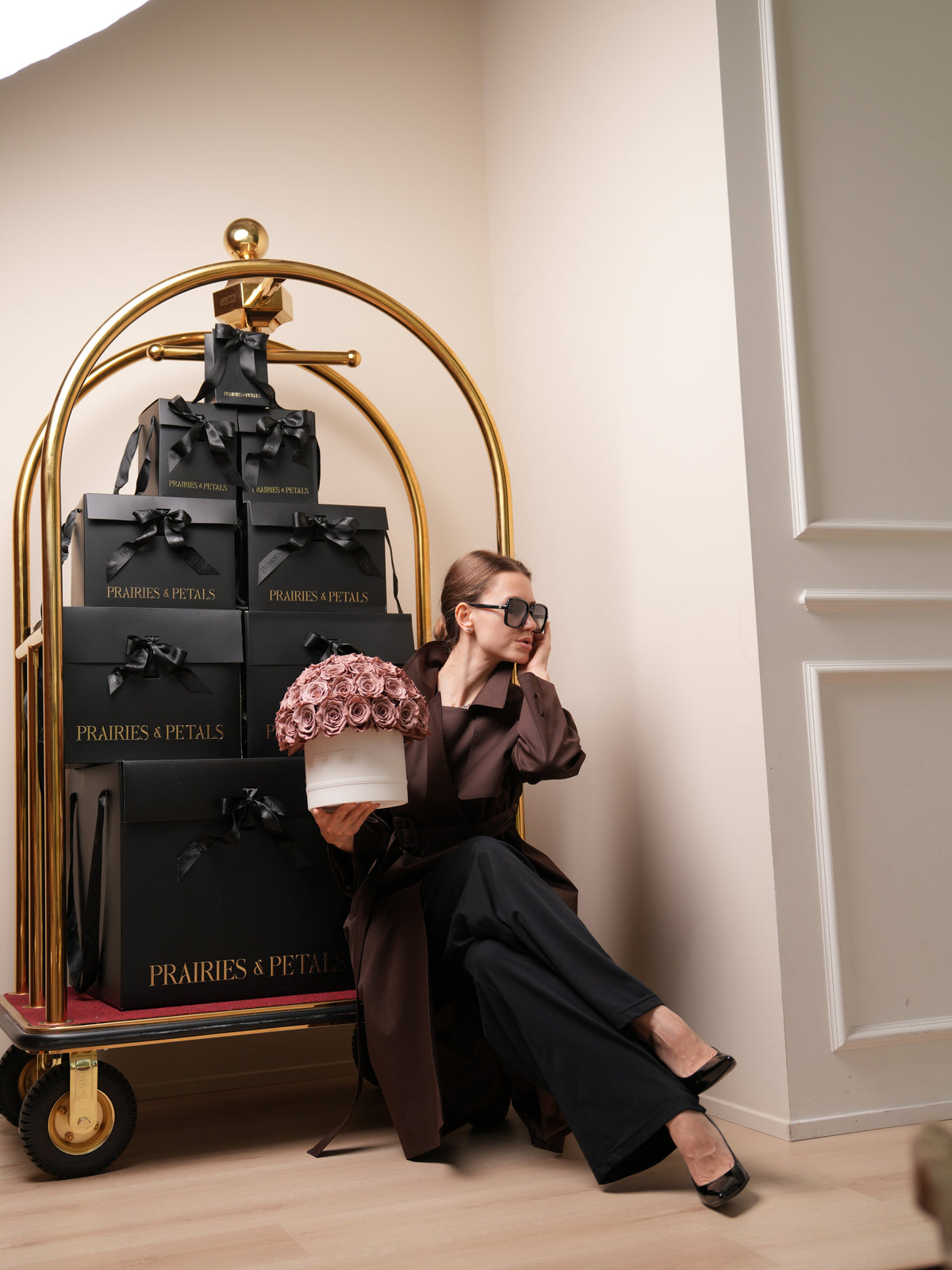 Prairies & Petals fall collection, woman in a brown trench holding a dusty rose arrangement beside a gold luggage cart of black Prairies & Petals gift boxes