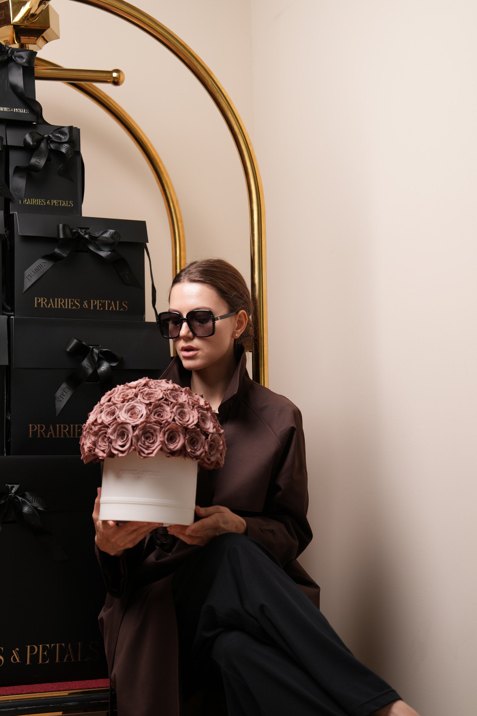 Prairies & Petals Fall Collection, model in a brown coat holding a mauve rose hatbox beside a gold bell cart of black gift boxes